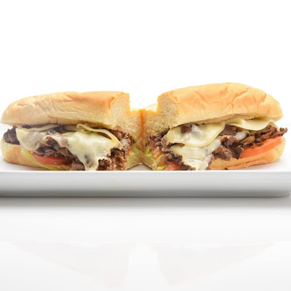 A Paisano's Pizza cheesesteak, cut in half, with melted cheese, steak, and tomatoes on a white plate.