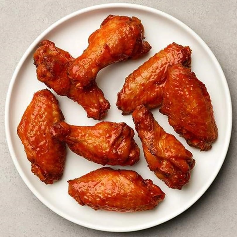 A white plate holds seven glossy, golden-brown chicken wings, coated in a sweet and savory sauce.